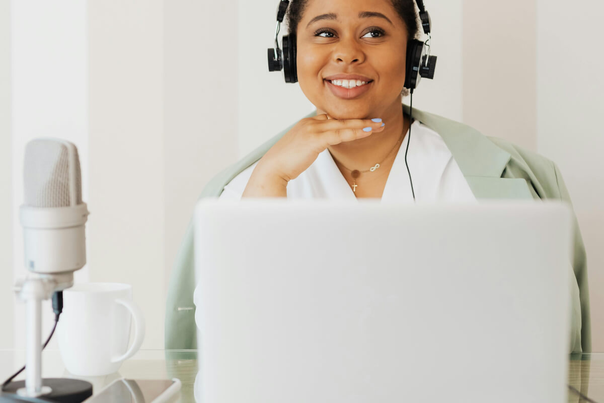 15 LinkedIn Profile Optimization Tips for Personal Branding 1 Smiling woman sits behind laptop with her hand below her chin, indicating she is framing her face. The figure is wearing headphones and there is a microphone next to the laptop, perhaps for podcasting or video creation.