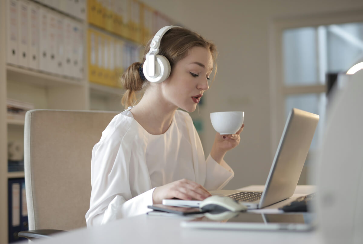 A photo of a woman in headphones drinking coffee and using her laptop