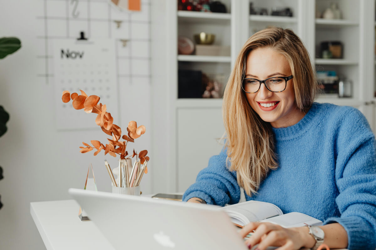 Blonde woman in blue top working happily on laptop with a plant, calendar, and shelves in the background